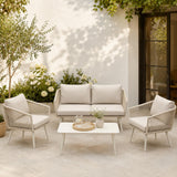 Outdoor patio set with beige sofa, chairs, and table on a patio with plants and a white wall.